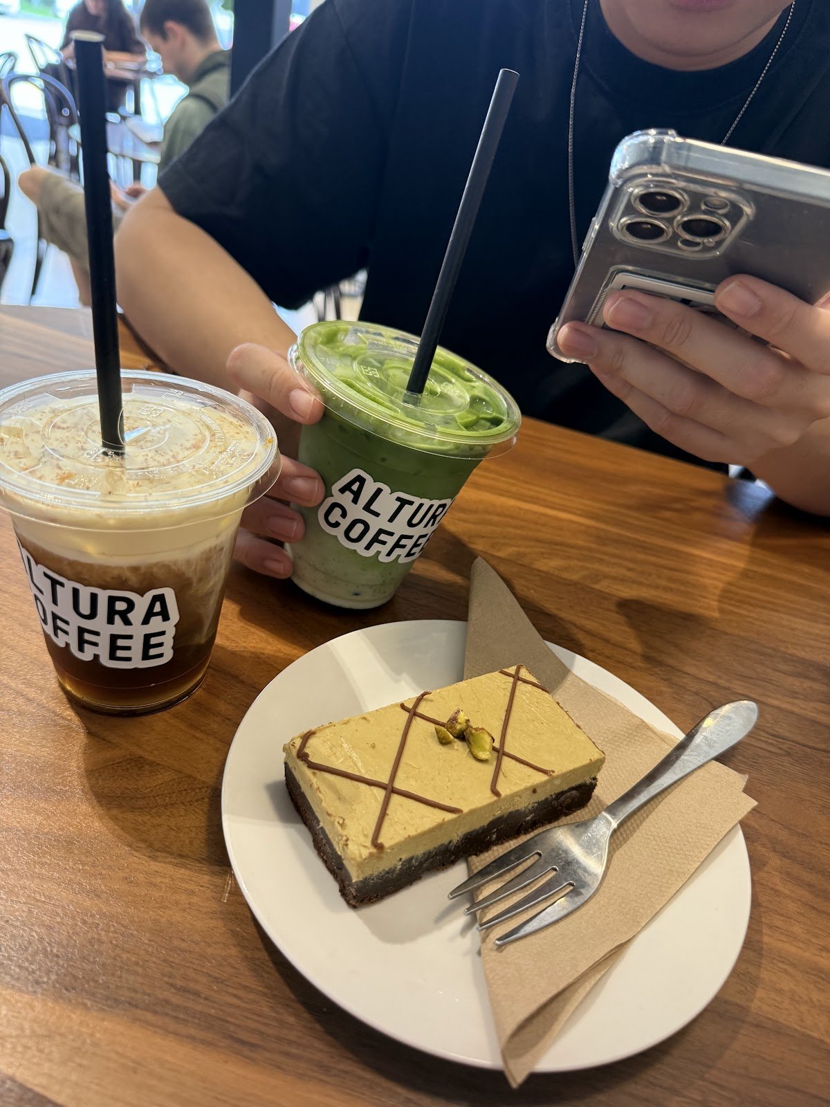 A close-up shot of an iced matcha latte in a clear plastic cup with black straw, served alongside another iced coffee drink on a wooden table at Alta Coffee, accompanied by a slice of layered cake topped with chocolate drizzle and pistachio garnish on a white plate.