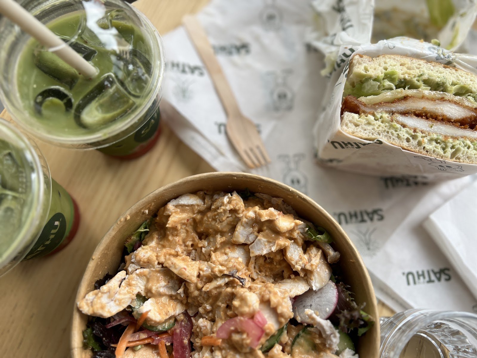 A top-down view of an iced matcha latte served in a clear plastic cup with ice cubes and two wooden straws, placed on a light wood table alongside another similar drink, a paper bowl containing a creamy chicken salad over greens, and half a sandwich wrapped in branded paper.