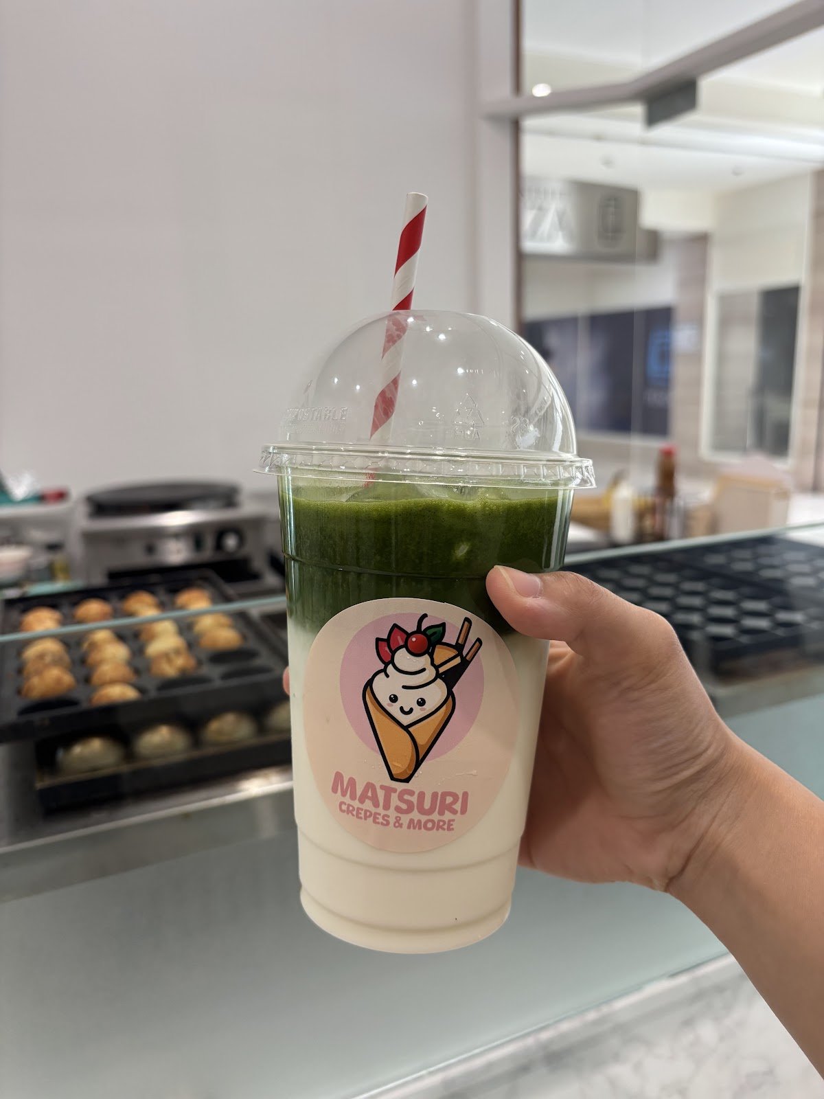 This is an iced matcha frappe served in a clear plastic cup with a dome lid and red-and-white striped straw, featuring a vibrant green matcha layer atop a creamy white base, decorated with a cartoon ice cream cone sticker.