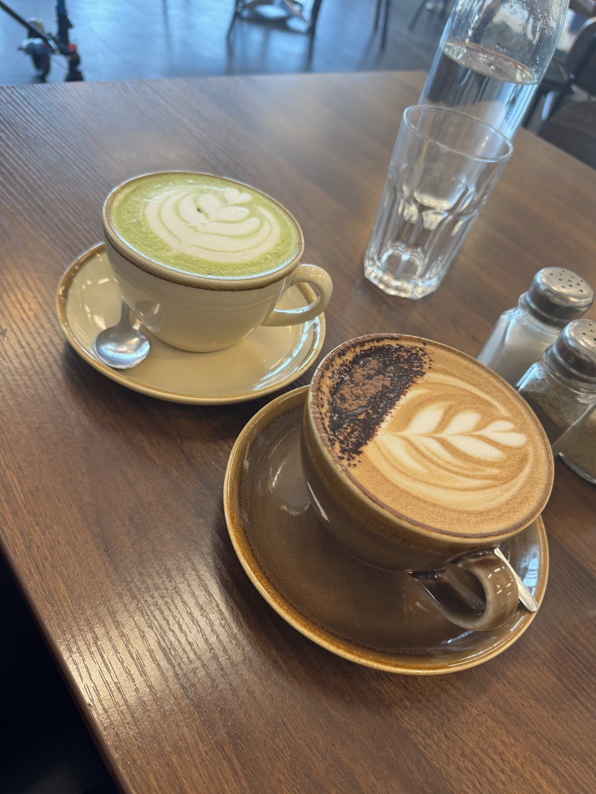 A wooden table displays two hot beverages: a light green matcha latte in an off-white ceramic cup with white leaf-patterned foam art on its surface, served alongside a brown frothy coffee drink topped with dark chocolate shavings and featuring similar leaf-shaped latte art inside both cups.