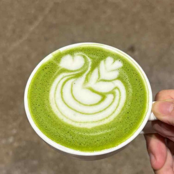 A hand holds a white ceramic cup containing hot matcha latte with intricate green tea foam art featuring swirling patterns in contrasting milk-colored froth against its vibrant emerald-green surface.