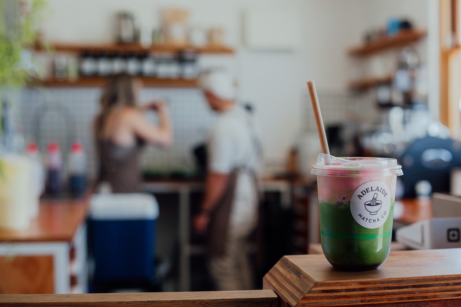 An iced matcha latte served in a clear plastic cup with a straw, featuring vibrant green liquid layered over pink foam at the top, presented on a wooden counter inside a bustling café background where baristas are working behind shelves stocked with jars and bottles.