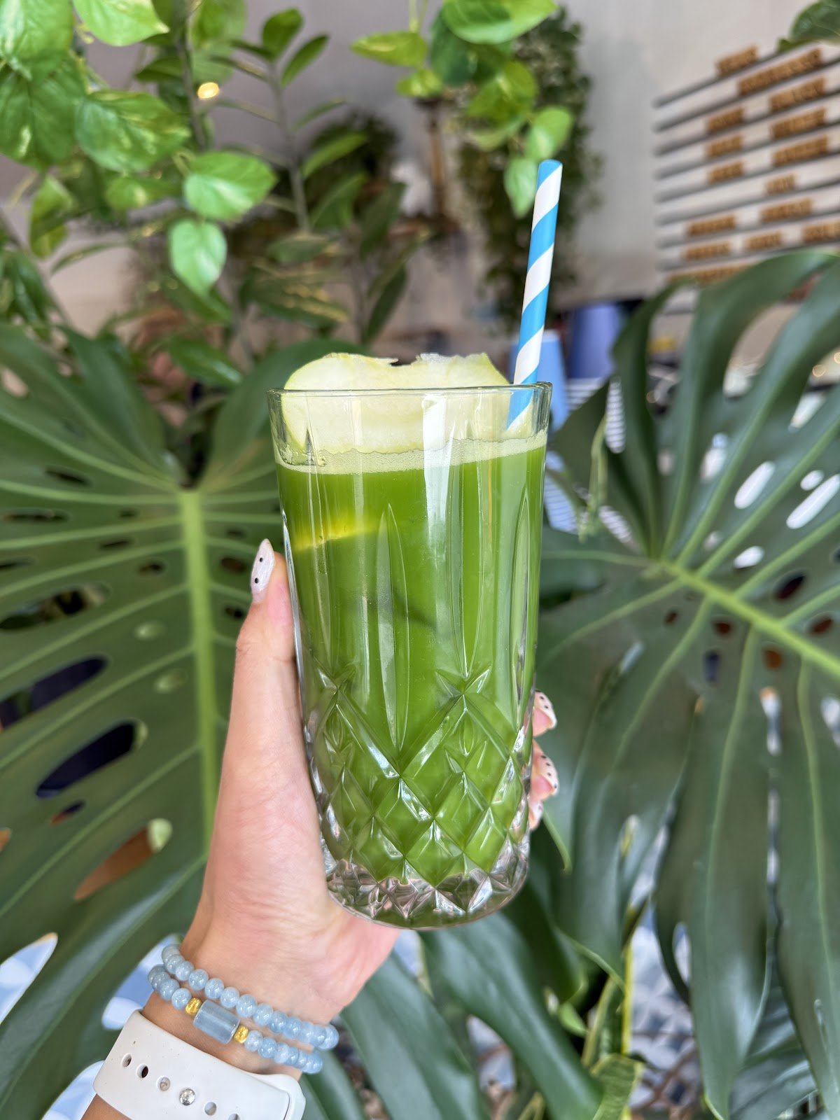 This is an iced green smoothie or juice served in a tall, faceted crystal glass, topped with a slice of apple and a blue-and-white striped straw, presented in a vibrant green hue against a backdrop of large monstera leaves.