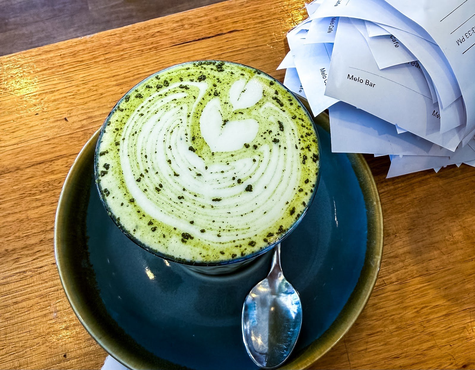 This is a hot matcha latte served in a ceramic cup on a matching saucer, featuring intricate white milk foam art shaped like a heart and swirls, dusted with fine green matcha powder, and accompanied by a spoon.