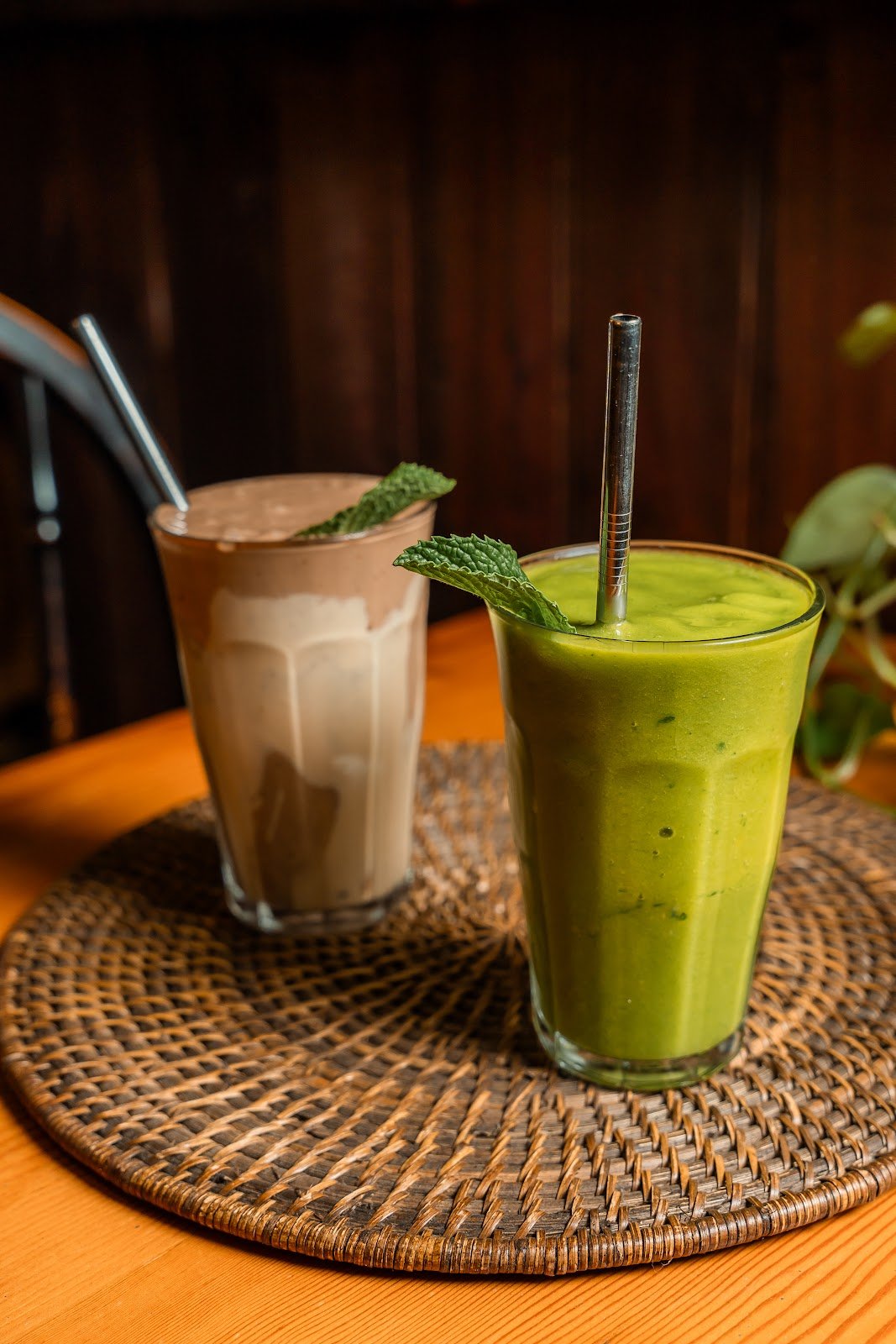 Two iced smoothies served in clear glasses on a woven placemat: one is a light brown chocolate milkshake topped with mint leaves, while the other is a vibrant green matcha latte also garnished with fresh mint; both feature metal straws and are presented against a dark wooden background.