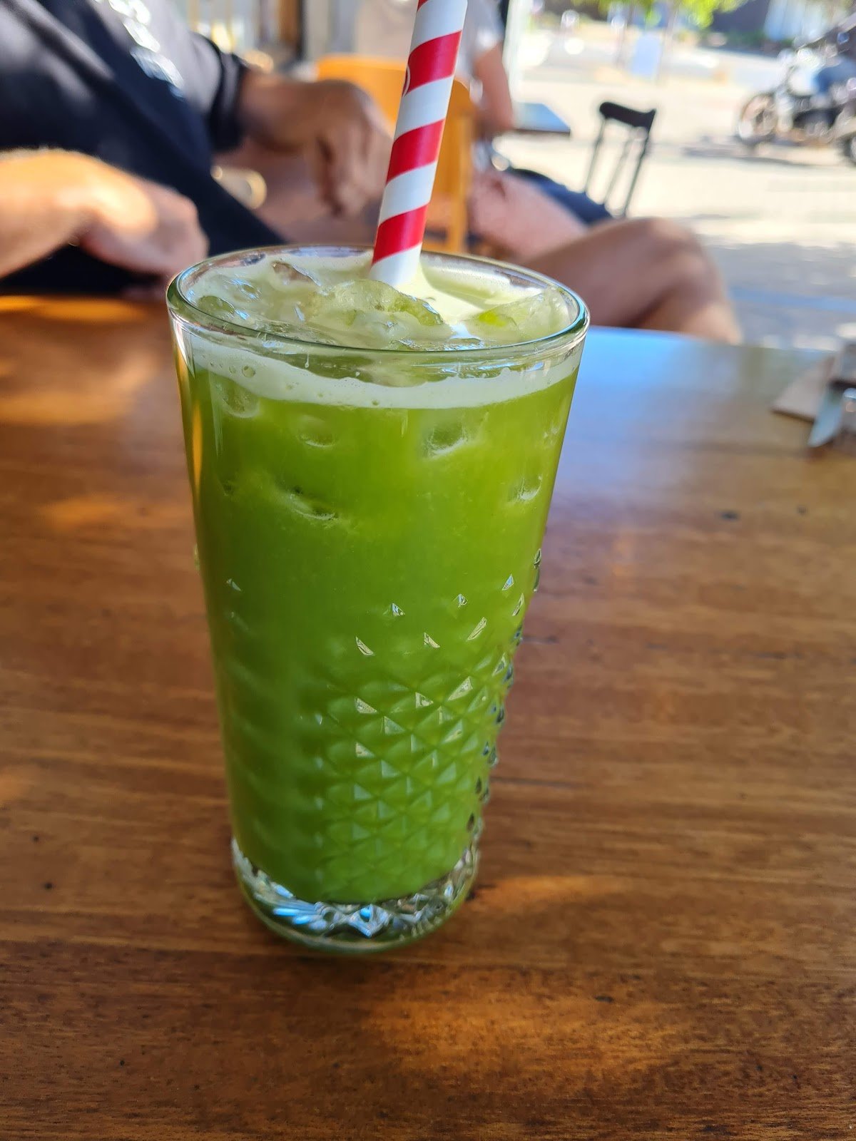 An iced matcha latte served in a textured clear glass with ice cubes and topped with whipped cream, featuring a red-and-white striped paper straw, presented on a wooden table outdoors under natural light.