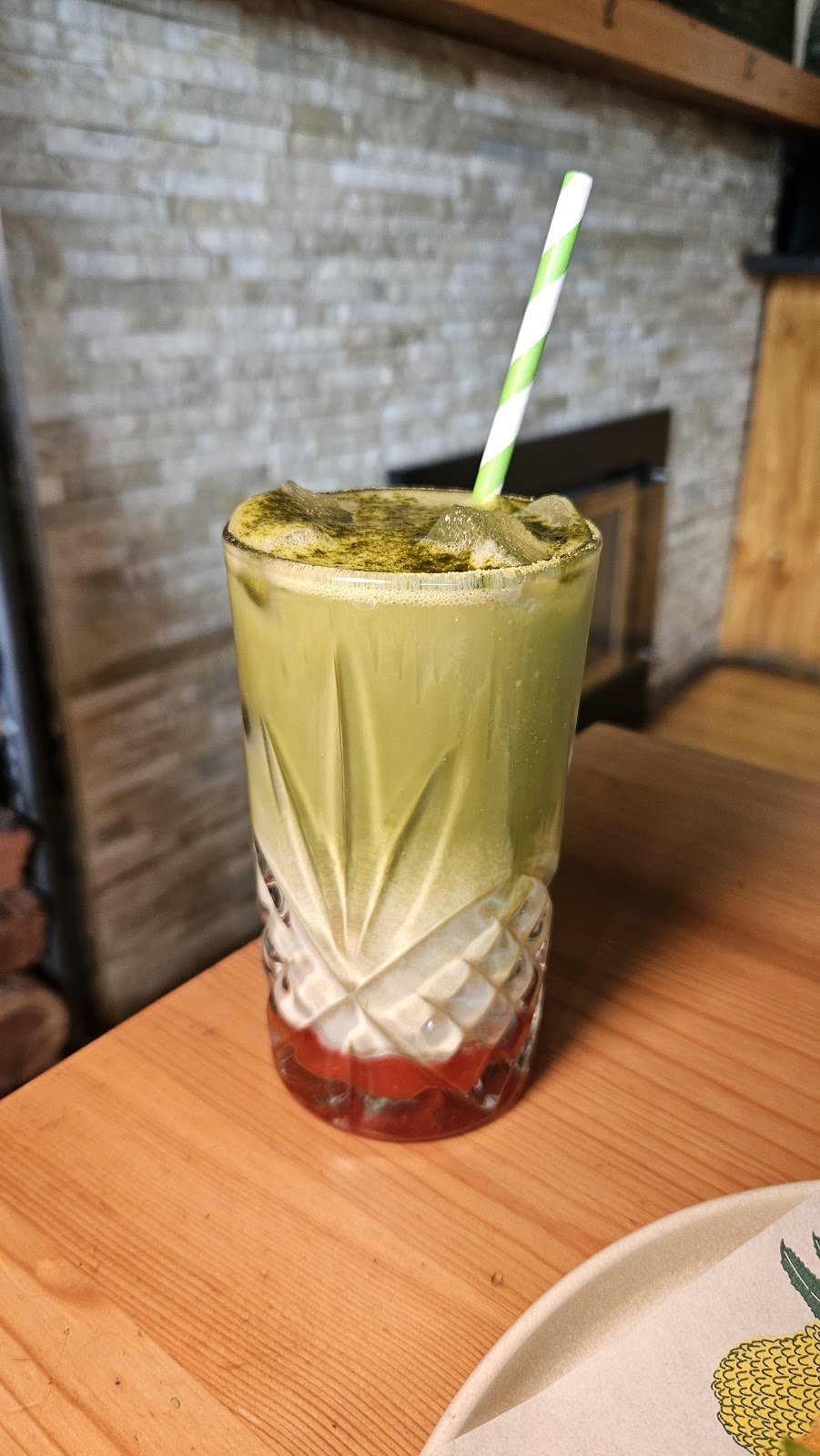 This is an iced matcha frappe served in a tall, patterned glass with a green-and-white striped straw, featuring a vibrant green matcha layer over a red syrup base and topped with a dusting of matcha powder.