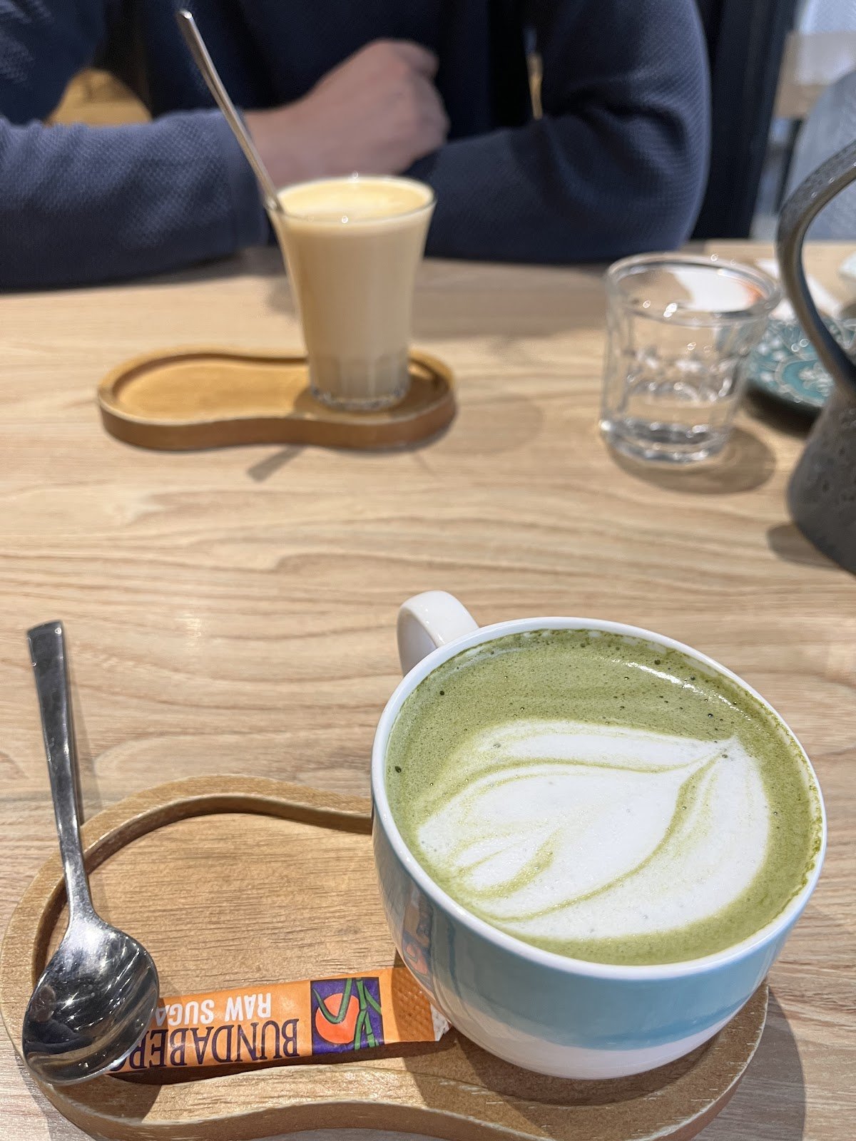 A close-up shot of a hot matcha latte served in a white ceramic cup with blue accents, featuring delicate leaf-shaped foam art on its green surface, resting on a wooden coaster alongside an orange Bundaberg raw sugar packet and spoon; another iced coffee drink sits further back on the same light wood table.