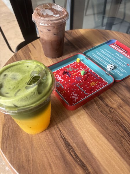 A wooden table displays two iced beverages in clear plastic cups—one with a layered green matcha top over orange liquid and another chocolate frappe topped with ice cream—alongside an open red tin containing a Snakes & Ladders game board, dice, and instructions.