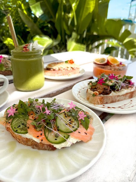 A refreshing iced matcha latte served in a clear mason jar with a gold straw, topped with whipped cream and edible flowers, presented alongside gourmet avocado toast on white plates at an outdoor table surrounded by lush greenery.