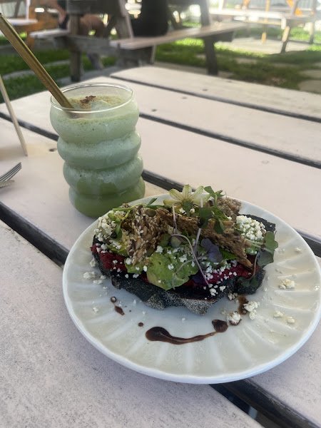 An iced matcha latte served in a ribbed clear plastic cup with a gold straw sits beside an open-faced avocado toast topped with crumbled cheese, microgreens, fried onions, and dark sauce drizzle on a white ceramic plate at an outdoor wooden picnic table setting.