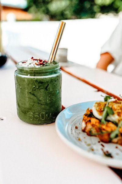 An iced matcha latte served in a clear mason jar with visible "530ML" embossing, topped with white coconut flakes and dark sprinkles, presented alongside a plated dish on an outdoor table setting.