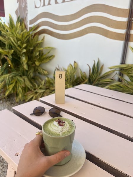A hand holds a light green ceramic cup containing an iced matcha latte topped with white foam and dried flower garnish on a pale pink outdoor wooden table beside sunglasses and a numbered sign.