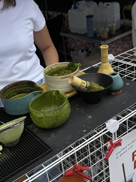A close-up of a traditional Japanese matcha preparation featuring vibrant green powdered tea whisked in ceramic bowls with bamboo chasen tools on a black countertop at an outdoor market stall displaying various pottery vessels and containers for beverages or food items.