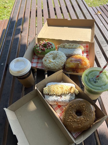 A matcha iced latte served in a clear plastic cup with green foam and red-and-white striped straw sits beside two open cardboard bakery boxes containing assorted pastries including glazed donuts, powdered sugar-covered treats, and cream-filled cakes on checkered paper liners, all arranged outdoors on wooden slats under natural daylight.