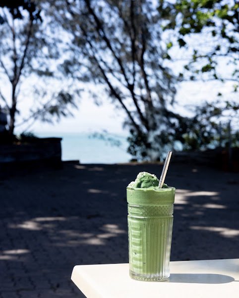 An iced matcha latte served in a ribbed clear glass with a metal straw, topped with fluffy green whipped cream, set on an outdoor white table against a backdrop of trees and ocean view under bright sunlight.
