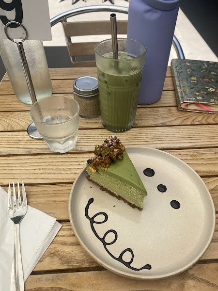 A slice of green matcha cheesecake with crumbly brown topping served on a smiley-face plate alongside an iced matcha latte in a clear glass with a metal straw, all arranged on a wooden table next to water glasses and a blue bottle.