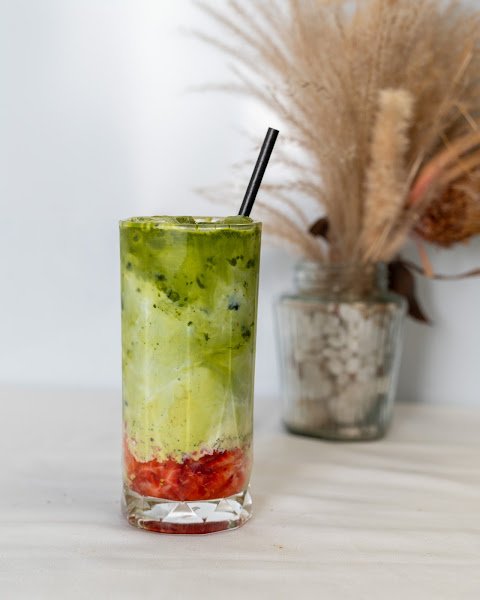 An iced matcha latte served in a tall clear glass with visible ice cubes and red fruit garnish at the bottom, featuring black straw stirrer against soft white background with dried pampas grass decoration nearby.