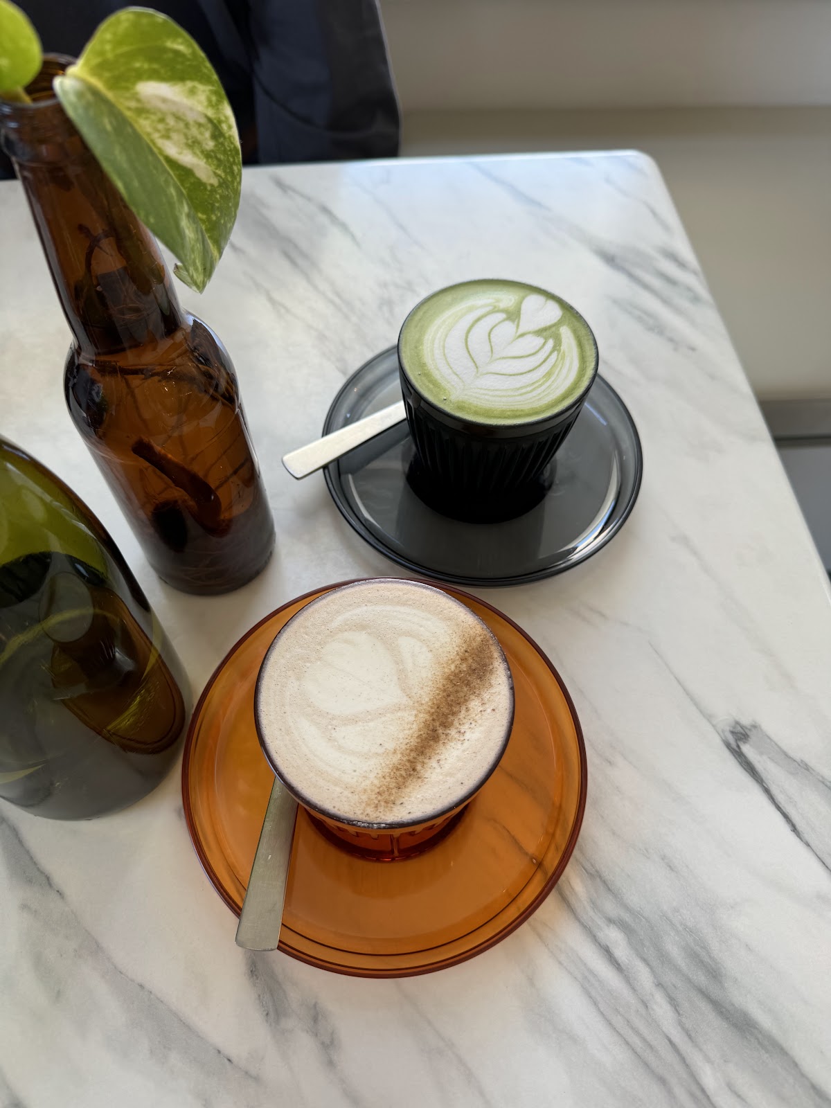 A hot matcha latte in a black cup with a delicate white leaf latte art design, served on a black saucer alongside a caramelized oat milk latte in an amber glass cup with a dusting of cinnamon, both resting on a white marble table with decorative brown glass bottles and a green leaf.