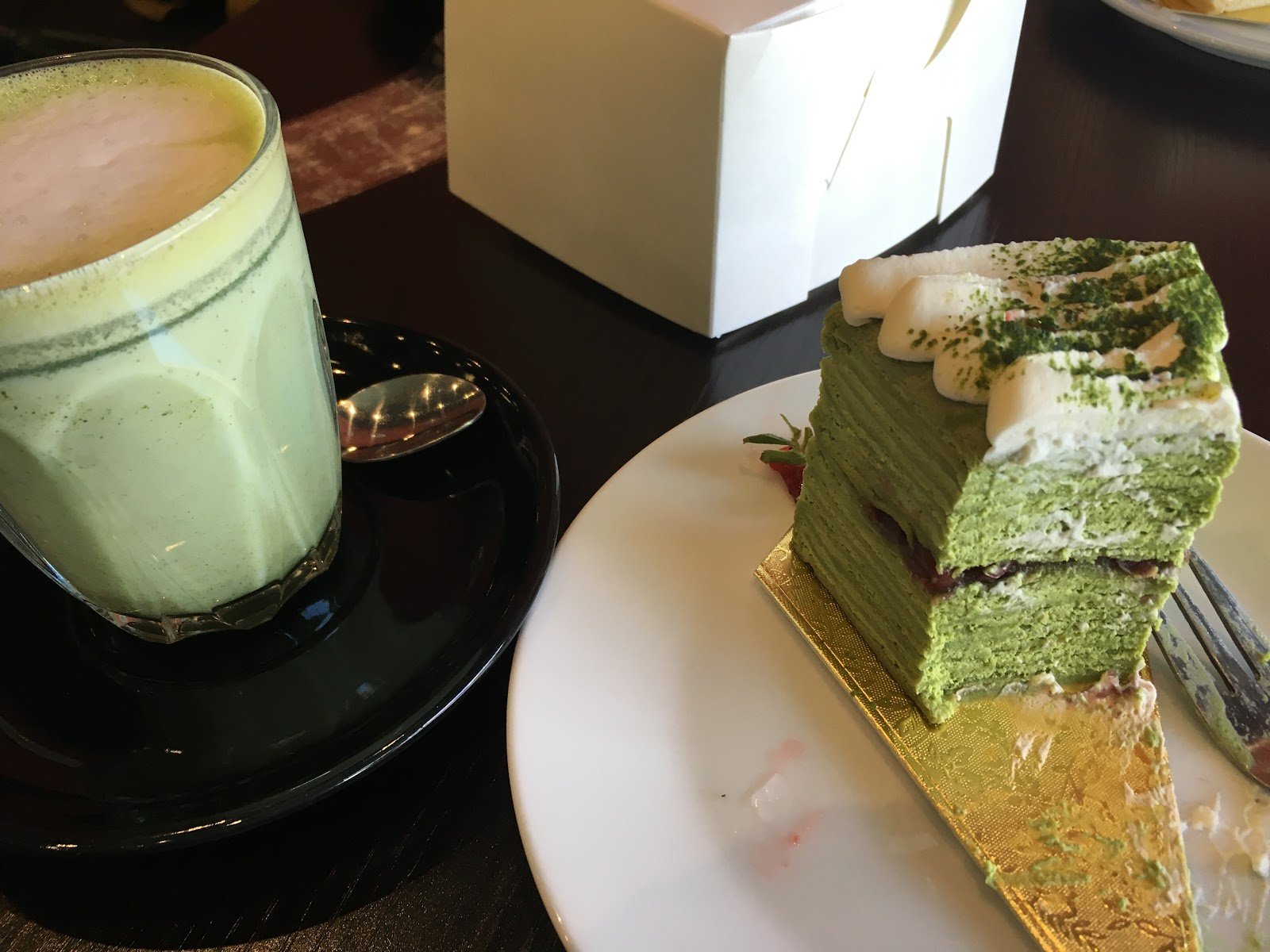 A vibrant green matcha latte in a clear glass cup sits beside a layered matcha cake with white cream and green powder garnish, both presented on a dark table with a white plate and gold foil base.