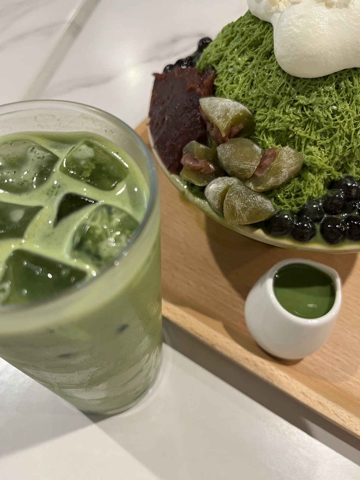 This is an iced matcha latte served in a clear glass with ice cubes, accompanied by a matcha-flavored shaved ice dessert in a bowl topped with whipped cream, black pearls, and candied grapefruit segments, all presented on a wooden tray with a small white cup of matcha sauce.