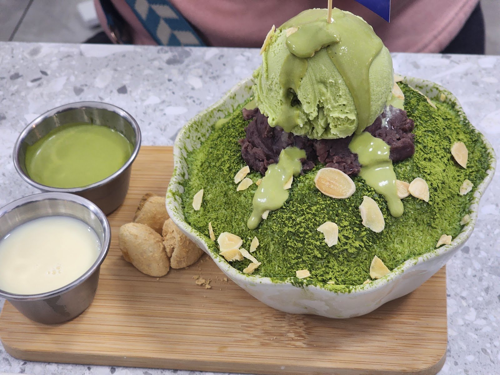 This is a vibrant, iced matcha shaved ice dessert served in a white ceramic bowl, topped with a scoop of green matcha ice cream, sliced almonds, dark red bean paste, and drizzled with matcha syrup, accompanied by two small metal cups of matcha and milk sauces, all presented on a wooden board.