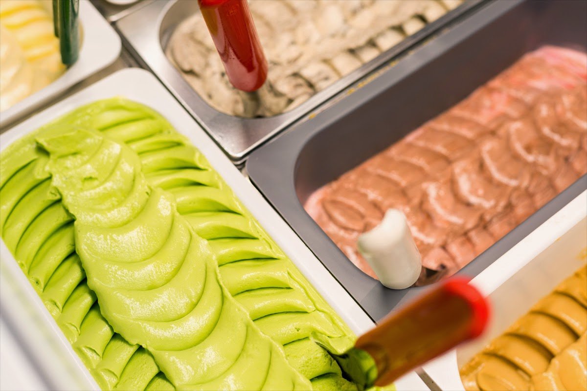 This image displays a close-up of freshly scooped matcha ice cream in a metal serving tray with decorative swirls on top, presented alongside other flavored options like chocolate and vanilla at an iced dessert counter without any cup or glass vessel visible for consumption.