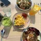 A top-down view of a light wood table featuring three bowls—one with chicken salad topped with drizzled sauce, another with noodles garnished by fried egg and shredded meat—and one plate holding golden french fries alongside ketchup; accompanied are two iced beverages in textured glasses (one green matcha latte frappe with cucumber slice, the other orange-colored drink), a water glass, sunglasses resting on a smartphone, all presented under bright natural daylight.