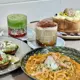 This vibrant photo displays a layered iced matcha frappe in a clear glass, topped with red fruit and green matcha foam, alongside a caramelized iced coffee in a textured glass, a creamy dessert cake with edible flowers and a chocolate drizzle, and a bowl of creamy pasta with a poached egg and herbs, all arranged on a wooden table with a white tiled background.