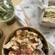 A top-down view of an iced matcha latte served in a clear plastic cup with ice cubes and two wooden straws, placed on a light wood table alongside another similar drink, a paper bowl containing a creamy chicken salad over greens, and half a sandwich wrapped in branded paper.
