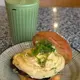 This is an iced matcha frappe served in a ribbed green glass with a white and gold striped straw, accompanied by a gourmet burger topped with fresh green onions and creamy sauce, both presented on a patterned plate against a speckled countertop.