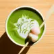 This is a hot matcha latte served in a white ceramic cup, featuring intricate white milk foam art of a stylized bird and garnished with a skewer of two small mochi (one pink, one white) resting on the surface.