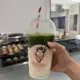 This is an iced matcha frappe served in a clear plastic cup with a dome lid and red-and-white striped straw, featuring a vibrant green matcha layer atop a creamy white base, decorated with a cartoon ice cream cone sticker.