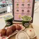 This photo displays two iced matcha lattes in clear plastic cups with lids and branded stickers, alongside a tray of golden-brown, sugar-dusted fried dough balls and two white mochi cakes—one topped with a red strawberry—presented on a wooden tray with red-and-white checkered paper, all set against a pink drink menu featuring various iced matcha and fruit-flavored beverages.