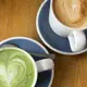Two white ceramic cups with blue saucers sit on a wooden table; one contains hot matcha latte featuring heart-shaped foam art in vibrant green, while the other holds an espresso-based drink topped with light brown milk foam also decorated with intricate leaf patterns.