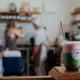 An iced matcha latte served in a clear plastic cup with a straw, featuring vibrant green liquid layered over pink foam at the top, presented on a wooden counter inside a bustling café background where baristas are working behind shelves stocked with jars and bottles.