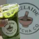 A close-up of an iced matcha latte served in a clear plastic cup with a wooden straw, featuring vibrant green coloration from frothy whipped cream on top and visible ice cubes inside; held by a hand against a blurred background displaying a circular logo that includes imagery of a bowl and whisk.