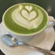 This is a hot matcha latte served in a white ceramic cup on a matching saucer, featuring intricate white milk foam art in the shape of two interlocking hearts on a vibrant green surface, accompanied by a silver spoon.