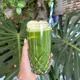 This is an iced green smoothie or juice served in a tall, faceted crystal glass, topped with a slice of apple and a blue-and-white striped straw, presented in a vibrant green hue against a backdrop of large monstera leaves.