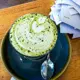 This is a hot matcha latte served in a ceramic cup on a matching saucer, featuring intricate white milk foam art shaped like a heart and swirls, dusted with fine green matcha powder, and accompanied by a spoon.