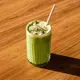 This is an iced matcha latte served in a textured green glass with a white straw, topped with a creamy white foam and sprinkled with matcha powder, presented with a warm, sunlit wooden background.