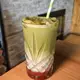 This is an iced matcha frappe served in a tall, patterned glass with a green-and-white striped straw, featuring a vibrant green matcha layer over a red syrup base and topped with a dusting of matcha powder.