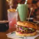 A vibrant green iced matcha frappe served in a tall, ribbed glass with a green-and-white striped straw and a decorative orange slice garnish, presented alongside a burger on a wooden board.