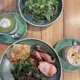 A top-down view of a wooden table featuring a hot matcha latte in a glass cup with a white heart-shaped latte art, a separate iced frappe in a glass topped with crumbled nuts, and a green bowl of fresh mixed greens salad, all presented in muted green ceramic vessels with a rustic, natural aesthetic.