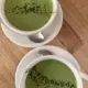 Two hot matcha lattes served in white ceramic cups on matching saucers, each topped with a sprinkle of loose matcha powder, presented in a vibrant green hue against a light wooden surface.