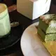 A vibrant green matcha latte in a clear glass cup sits beside a layered matcha cake with white cream and green powder garnish, both presented on a dark table with a white plate and gold foil base.