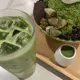 This is an iced matcha latte served in a clear glass with ice cubes, accompanied by a matcha-flavored shaved ice dessert in a bowl topped with whipped cream, black pearls, and candied grapefruit segments, all presented on a wooden tray with a small white cup of matcha sauce.