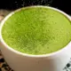 A close-up of a hot matcha latte served in an off-white ceramic cup with a handle, topped with fine green powder dusting over its frothy surface against a speckled countertop background.