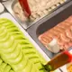 This image displays a close-up of freshly scooped matcha ice cream in a metal serving tray with decorative swirls on top, presented alongside other flavored options like chocolate and vanilla at an iced dessert counter without any cup or glass vessel visible for consumption.