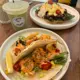 An iced matcha latte in a clear plastic cup with a white lid and straw, featuring a creamy green beverage topped with a small white circular logo, sits beside a plate of shrimp tacos and a plate of eggs benedict on a wooden table.